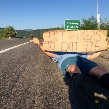 Mountain Man laying on the shoulder of a road and holding a sign trying to hitchhike