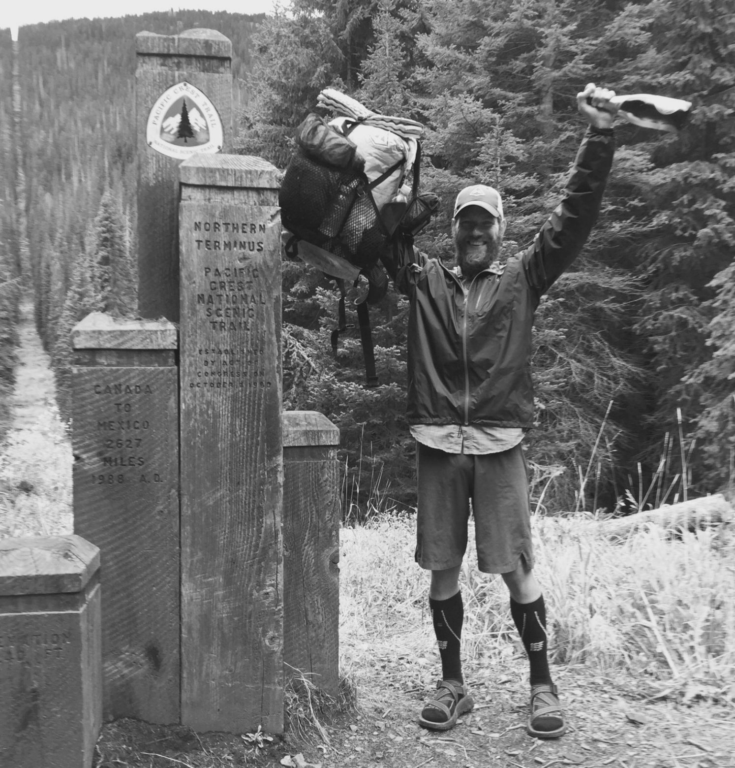 Mountain Man holding backpack and a bottle of champagne at the PCT northern terminus